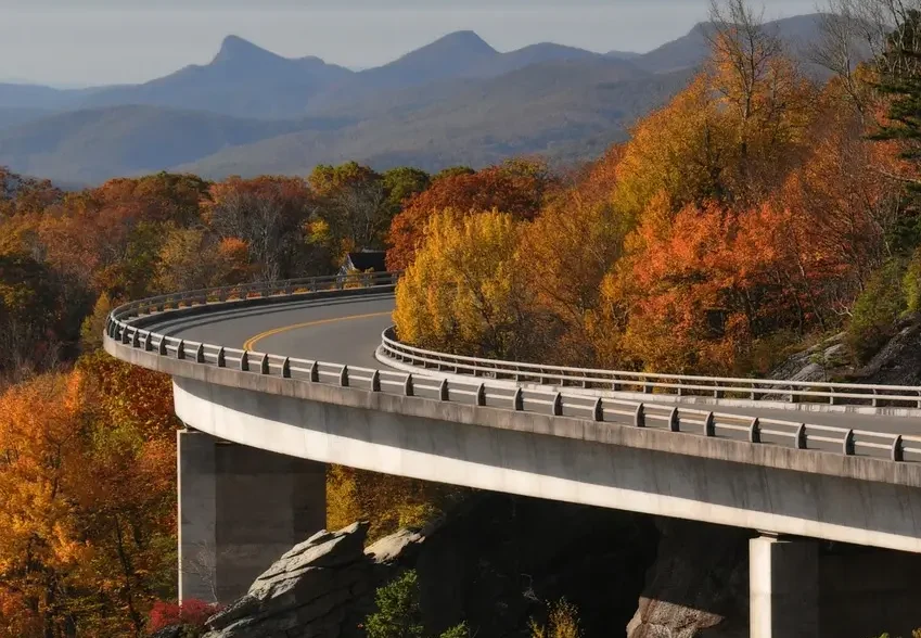 A curved bridge on the Blue Ridge Parkway cuts through a vibrant fall forest, with colorful autumn leaves in shades of orange, yellow, and red. The mountains in the background rise softly, creating a serene and majestic view, perfect for a peaceful road trip through nature.